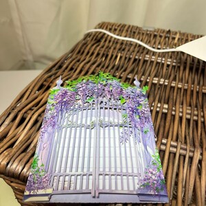 May include: A decorative card featuring a purple gate adorned with wisteria flowers. The gate is detailed with vertical bars and is surrounded by cascading purple blossoms. The card is set against a woven basket background, creating a charming, floral theme.