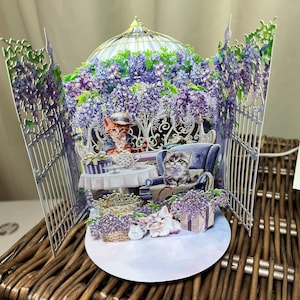May include: A 3D pop-up card featuring a whimsical scene with cats. The card depicts a garden with purple wisteria, a table, and a blue armchair. Two cats are seated, and two more are at the base of the card.