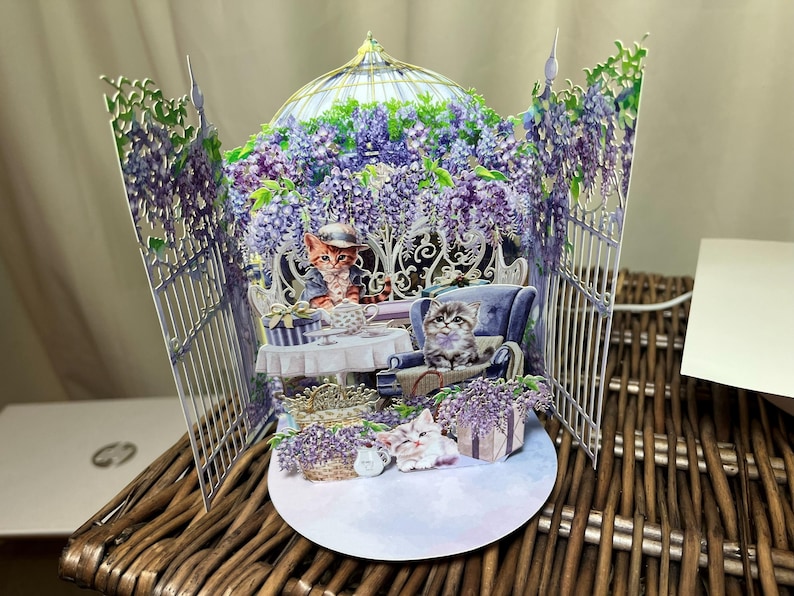 May include: A 3D pop-up card featuring a whimsical scene with cats. The card depicts a garden with purple wisteria, a table, and a blue armchair. Two cats are seated, and two more are at the base of the card.