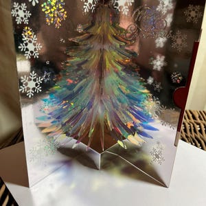 May include: A silver pop-up Christmas card with a holographic Christmas tree and snowflakes. The card is open, displaying the 3D tree design. The words "Merry Christmas" are visible on the card.