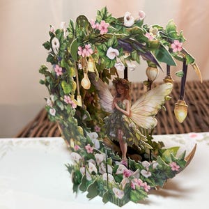 Może przedstawiać: Szczegółowa karta 3D pop-up przedstawiająca wróżkę w kwiatowej scenerii. Wróżka ma duże skrzydła i jest otoczona zielonymi liśćmi, różowymi kwiatami i wiszącymi lampionami. Karta jest umieszczona na neutralnym tle.