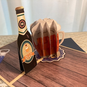 May include: A 3D pop-up card featuring a brown beer bottle with a white label that says "Beer" and a foam-topped beer mug with a brown handle. The card is on a wooden surface with a blue bottle opener.