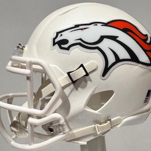 Könnte beinhalten: Weißer American-Football-Helm mit dem Logo der Denver Broncos. Das Logo ist ein weißer Pferdekopf mit schwarzen und orangefarbenen Akzenten. Der Helm hat ein weißes Gesichtsgitter und einen Kinnriemen. Ein Aufkleber mit der amerikanischen Flagge ist sichtbar.