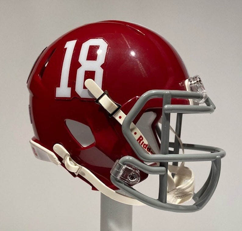 Puede incluir: Un casco de f&uacute;tbol americano rojo en miniatura con el n&uacute;mero "18" blanco en el lateral. El casco tiene una m&aacute;scara facial gris y correas blancas.