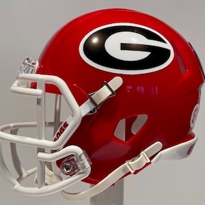 May include: A miniature red and white football helmet with a white face mask and a black and white "G" logo. The helmet has a white strap and buckle on the side.