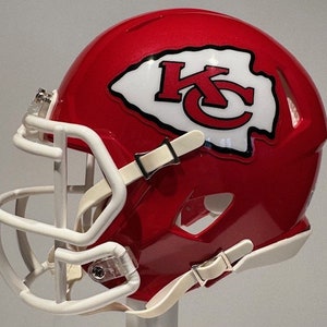 Puede incluir: Un casco de fútbol americano rojo y blanco con una máscara facial blanca y un logotipo "KC" blanco en el lateral. El casco es una versión en miniatura de un casco de fútbol americano profesional.