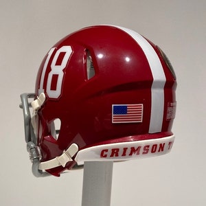 Puede incluir: Un casco de f&uacute;tbol americano en miniatura de color rojo y blanco con el n&uacute;mero "18" blanco en el lateral, una peque&ntilde;a pegatina de la bandera estadounidense y las palabras "CRIMSON TIDE" en la parte inferior.