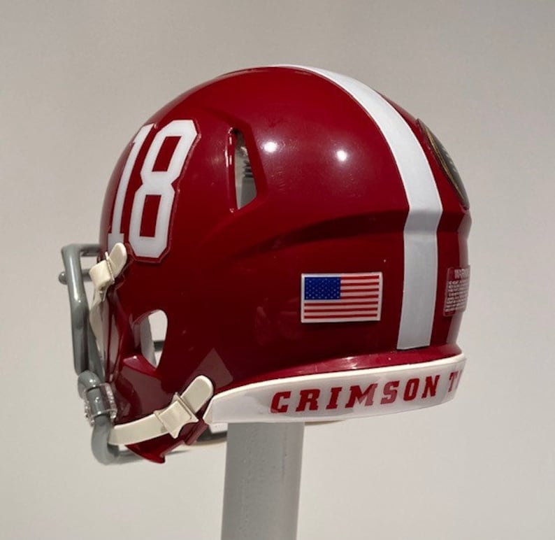 Puede incluir: Un casco de f&uacute;tbol americano en miniatura de color rojo y blanco con el n&uacute;mero "18" blanco en el lateral, una peque&ntilde;a pegatina de la bandera estadounidense y las palabras "CRIMSON TIDE" en la parte inferior.