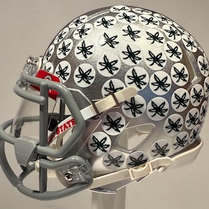 May include: A silver football helmet with a black and white Ohio State logo repeated on the surface. The helmet has a gray face mask and a white strap.