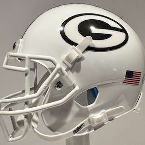 Puede incluir: Casco de fútbol americano blanco con un logotipo "G" negro y verde en el lateral. El casco tiene una máscara facial blanca y una pequeña pegatina de la bandera estadounidense en la parte posterior. El casco está hecho por Schutt.
