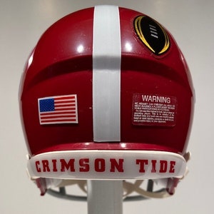 Puede incluir: Un casco de f&uacute;tbol rojo y blanco con una franja blanca en el centro. El casco tiene una peque&ntilde;a pegatina de la bandera estadounidense en el lateral y una etiqueta de advertencia en la parte posterior. El casco tiene una banda blanca alrededor de la parte inferior con las palabras "CRIMSON TIDE" en letras negras.