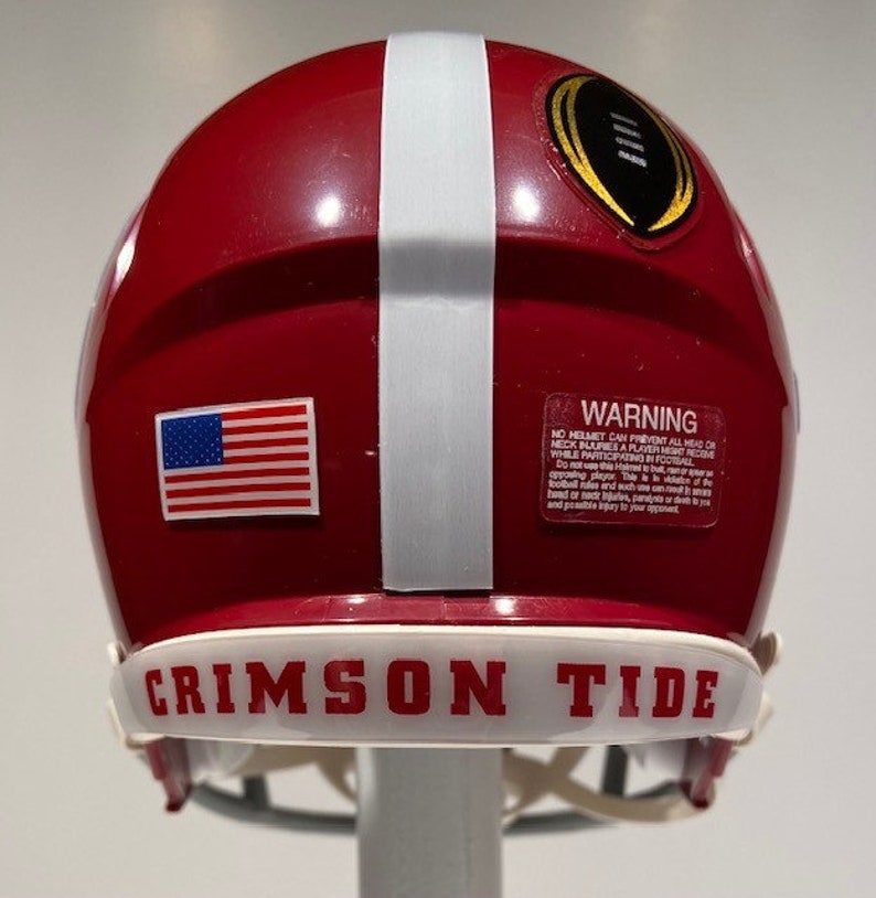 Puede incluir: Un casco de f&uacute;tbol rojo y blanco con una franja blanca en el centro. El casco tiene una peque&ntilde;a pegatina de la bandera estadounidense en el lateral y una etiqueta de advertencia en la parte posterior. El casco tiene una banda blanca alrededor de la parte inferior con las palabras "CRIMSON TIDE" en letras negras.