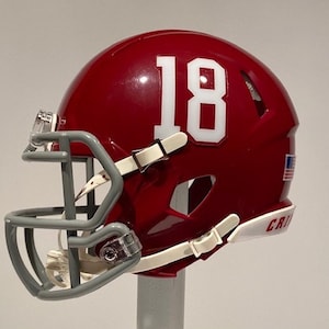 Puede incluir: Un casco de f&uacute;tbol americano rojo y blanco con una m&aacute;scara facial gris. El casco tiene el n&uacute;mero "18" en el lateral y la palabra "CRIS" en la parte inferior.