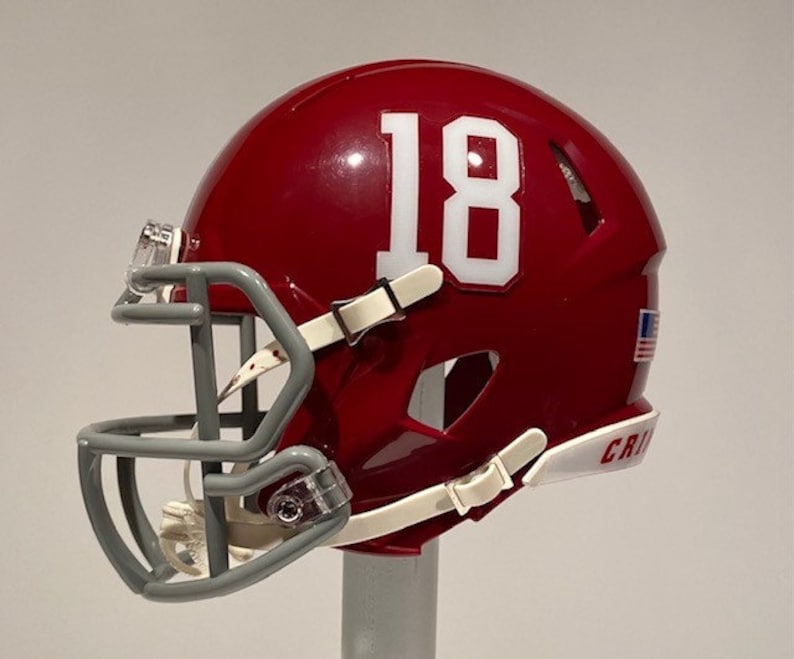 Puede incluir: Un casco de f&uacute;tbol americano rojo y blanco con una m&aacute;scara facial gris. El casco tiene el n&uacute;mero "18" en el lateral y la palabra "CRIS" en la parte inferior.