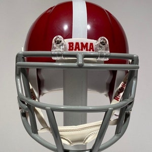 Puede incluir: Un casco de f&uacute;tbol americano rojo y blanco con una m&aacute;scara facial gris. El casco tiene la palabra "BAMA" escrita en la parte delantera. El casco est&aacute; hecho por Riddell.