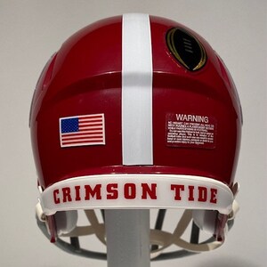 Puede incluir: Un casco de f&uacute;tbol americano rojo y blanco con una franja blanca a un lado. El casco tiene una peque&ntilde;a pegatina de la bandera estadounidense en un lado y un logotipo dorado de campeonato de f&uacute;tbol en la parte posterior. El casco tiene una banda blanca alrededor de la parte inferior con las palabras "Crimson Tide" en letras rojas.