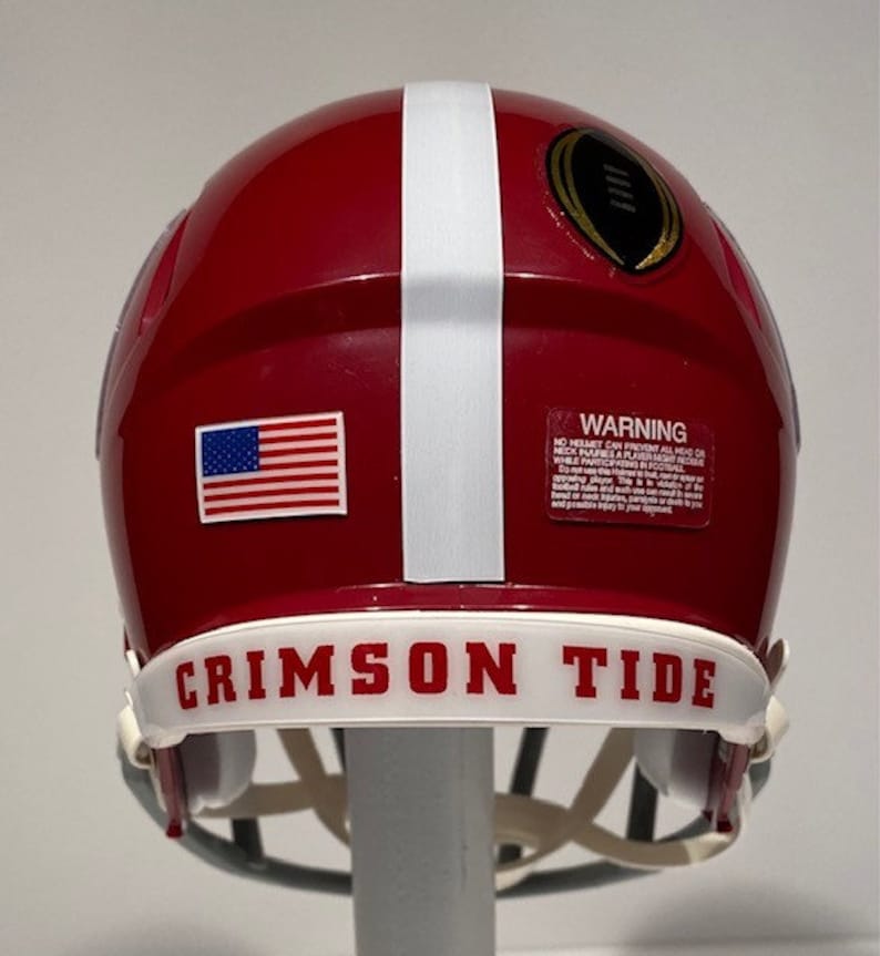 Puede incluir: Un casco de f&uacute;tbol americano rojo y blanco con una franja blanca a un lado. El casco tiene una peque&ntilde;a pegatina de la bandera estadounidense en un lado y un logotipo dorado de campeonato de f&uacute;tbol en la parte posterior. El casco tiene una banda blanca alrededor de la parte inferior con las palabras "Crimson Tide" en letras rojas.