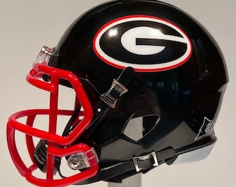 May include: A black and white football helmet with a red face mask. The helmet has a white "G" logo on a black oval with a red outline. The helmet is a miniature replica.