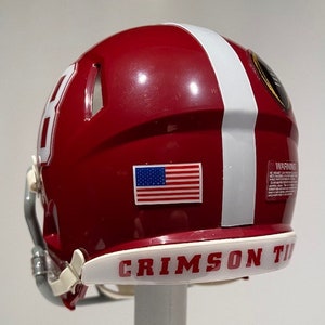 Puede incluir: Un casco de f&uacute;tbol americano rojo y blanco con una peque&ntilde;a pegatina de la bandera estadounidense en el lateral. El casco tiene una franja blanca en el lateral y las palabras "CRIMSON TIDE" en la parte inferior.