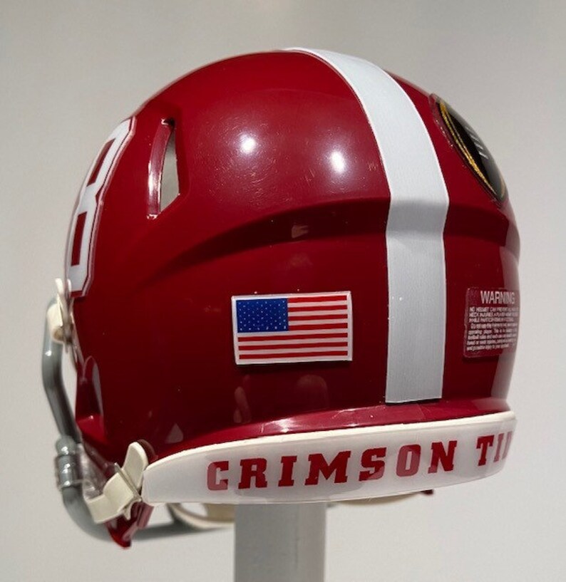 Puede incluir: Un casco de f&uacute;tbol americano rojo y blanco con una peque&ntilde;a pegatina de la bandera estadounidense en el lateral. El casco tiene una franja blanca en el lateral y las palabras "CRIMSON TIDE" en la parte inferior.