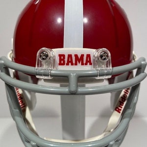 Puede incluir: Un casco de f&uacute;tbol americano en miniatura de color rojo y blanco con una m&aacute;scara facial gris. El casco tiene una franja blanca en el centro y una pegatina blanca con la palabra "BAMA" en la parte delantera. El casco est&aacute; hecho por Riddell.