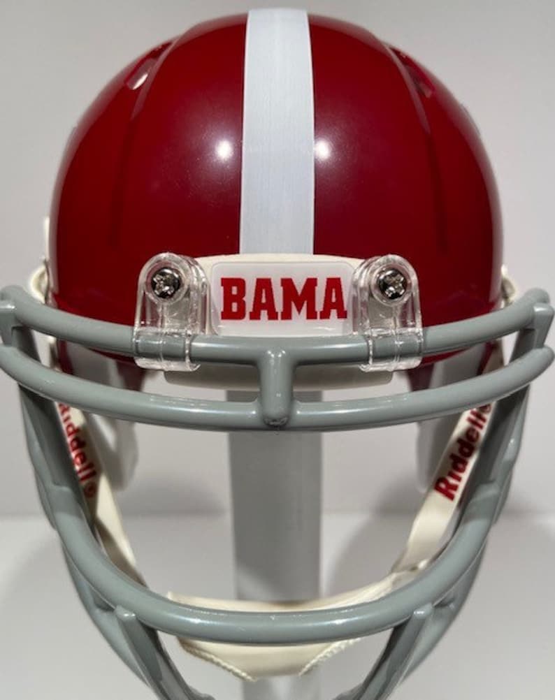 Puede incluir: Un casco de f&uacute;tbol americano en miniatura de color rojo y blanco con una m&aacute;scara facial gris. El casco tiene una franja blanca en el centro y una pegatina blanca con la palabra "BAMA" en la parte delantera. El casco est&aacute; hecho por Riddell.