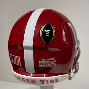 Puede incluir: Un casco de f&uacute;tbol miniatura rojo y blanco con un logotipo de Campeonato Nacional de la NCAA en oro y negro. El casco tiene una franja blanca en el lateral y las palabras "Crimson Tide" en la parte inferior.