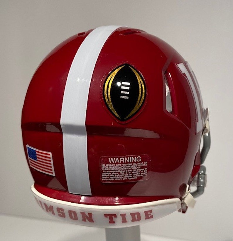 Puede incluir: Un casco de f&uacute;tbol miniatura rojo y blanco con un logotipo de Campeonato Nacional de la NCAA en oro y negro. El casco tiene una franja blanca en el lateral y las palabras "Crimson Tide" en la parte inferior.