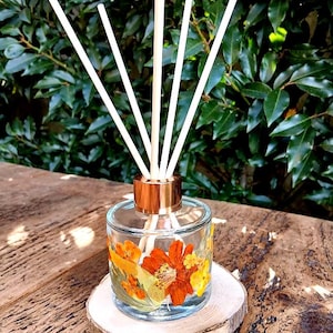 May include: A clear glass reed diffuser bottle with a metallic copper-coloured cap, featuring pressed orange and yellow flowers on the exterior. Five white reed sticks are inserted into the bottle, which rests on a circular wooden slice base.
