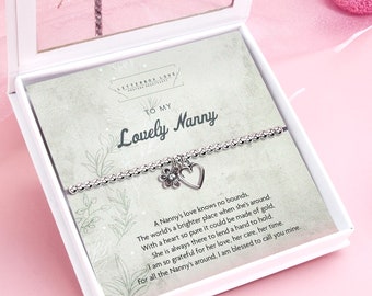 May include: A silver bracelet with a heart and flower charm, displayed in a white gift box. The card inside reads "To My Lovely Nanny" with a poem about a nanny's love. Pink Christmas baubles and decorations are in the background.
