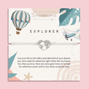 Puede incluir: Una pulsera de cordón blanco con un dije plateado en forma de corazón con un diseño de mapa del mundo. La tarjeta detrás de la pulsera tiene la palabra "EXPLORER" y una ilustración de globo aerostático. La tarjeta también tiene el texto "LETTERBOX LOVE" y "POSTIVE POSITIVITY".