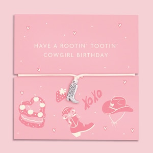 May include: A silver-toned cowboy boot charm on a thin white cord, attached to a pink card featuring white illustrations of a heart-shaped cake, a strawberry, a boot with a bow, a cowboy hat, and the text HAVE A ROOTIN' TOOTIN' COWGIRL BIRTHDAY.