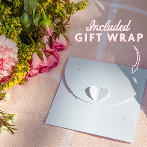 May include: A white gift wrap with a heart-shaped closure and small black dots. The text "Included GIFT WRAP" is in pink script. A bouquet of pink roses and carnations rests on a pink blanket.