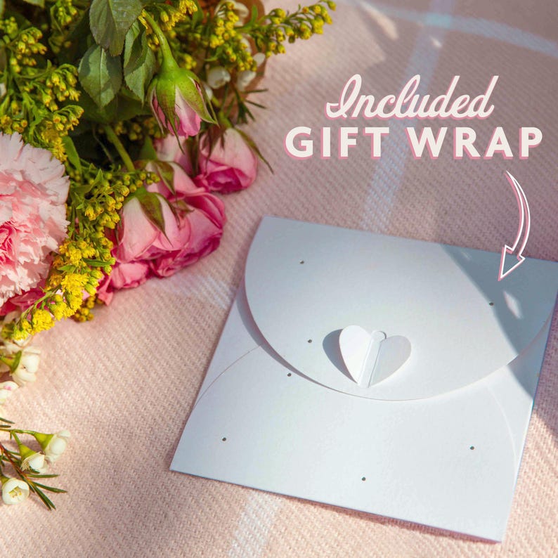 May include: A white gift wrap with a heart-shaped closure and small black dots. The text "Included GIFT WRAP" is in pink script. A bouquet of pink roses and carnations rests on a pink blanket.