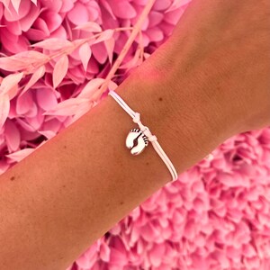 May include: A white string bracelet with a silver charm shaped like baby feet. The bracelet is tied on a wrist, set against a backdrop of pink flowers and foliage.