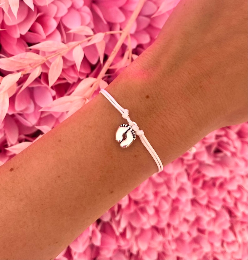 May include: A white string bracelet with a silver charm shaped like baby feet. The bracelet is tied on a wrist, set against a backdrop of pink flowers and foliage.