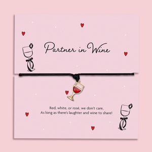 May include: A black cord bracelet with a gold-tone wine glass charm featuring red enamel. The bracelet is attached to a light pink card printed with the text Partner in Wine, Red, white, or rosé, we don't care, As long as there's laughter and wine to share!
