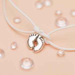 May include: A silver-colored charm in the shape of baby footprints is suspended on a white cord. The charm is the focal point, set against a soft pink background with scattered clear, faceted beads. The cord is knotted above the charm.