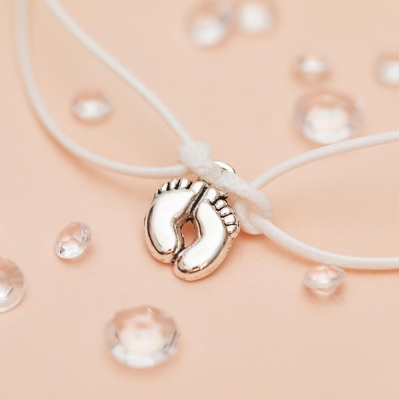 May include: A silver-colored charm in the shape of baby footprints is suspended on a white cord. The charm is the focal point, set against a soft pink background with scattered clear, faceted beads. The cord is knotted above the charm.