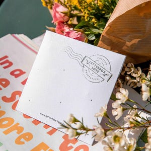May include: A square white card with small holes for jewelry, featuring a black circular stamp design that reads LETTERBOX LOVE POSTING POSITIVITY and the website www.letterbox-love.co printed at the bottom, surrounded by yellow and pink flowers.