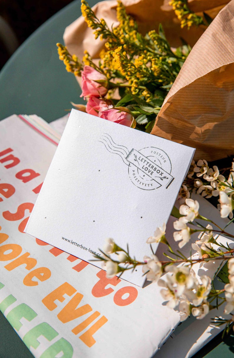 May include: A square white card with small holes for jewelry, featuring a black circular stamp design that reads LETTERBOX LOVE POSTING POSITIVITY and the website www.letterbox-love.co printed at the bottom, surrounded by yellow and pink flowers.