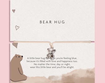 May include: A silver-toned bear charm on a light-colored string bracelet attached to a white card. The card features the text BEAR HUG, a brown bear illustration, and a poem about feeling blue, love, and happiness, decorated with small hearts.