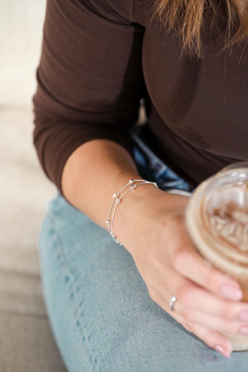 May include: A delicate silver bracelet with two strands, adorned with small, round beads and star-shaped charms. The bracelet is worn on a wrist, showcasing its fine details and elegant design. The jewelry is a subtle accessory.