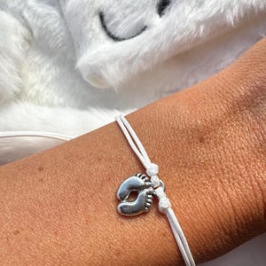 May include: A white cord bracelet featuring a silver-toned charm shaped like a pair of baby feet. The bracelet is secured with a simple knot mechanism, allowing for an adjustable fit around the wrist, showcasing a clean and minimalist design.