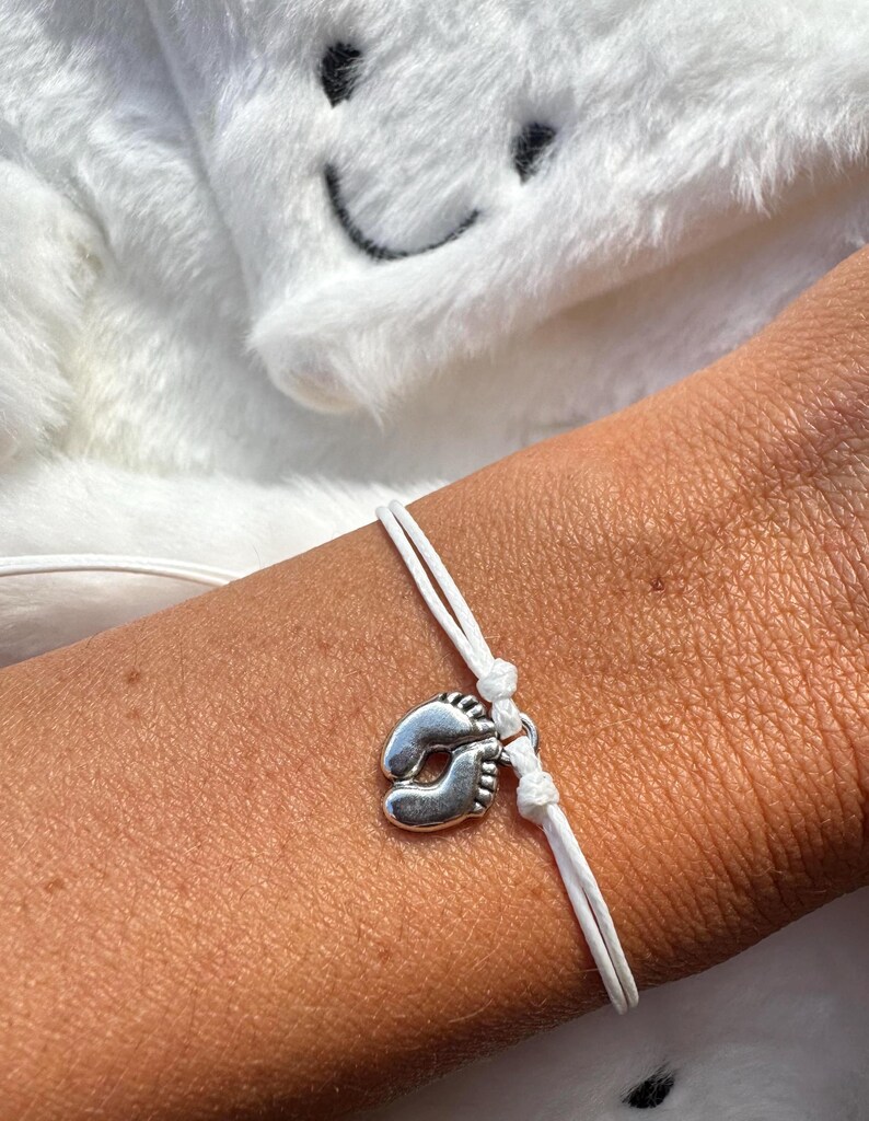 May include: A white cord bracelet featuring a silver-toned charm shaped like a pair of baby feet. The bracelet is secured with a simple knot mechanism, allowing for an adjustable fit around the wrist, showcasing a clean and minimalist design.