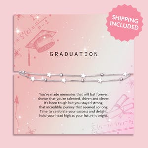 May include: A silver necklace with star charms is displayed on a pink graduation card. The card has the word "GRADUATION" at the top and a message below. A pink sticker with the words "SHIPPING INCLUDED" is in the top right corner.