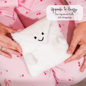 May include: A white, fluffy gift wrap with a smiling face, black eyes, and a curved mouth. The text "Upgrade to Hugsy" and "Our Signature Fluffy Gift Wrapping" is visible. The background is pink pajamas with a cherry pattern.