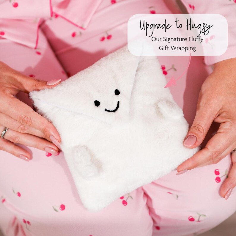 May include: A white, fluffy gift wrap with a smiling face, black eyes, and a curved mouth. The text "Upgrade to Hugsy" and "Our Signature Fluffy Gift Wrapping" is visible. The background is pink pajamas with a cherry pattern.