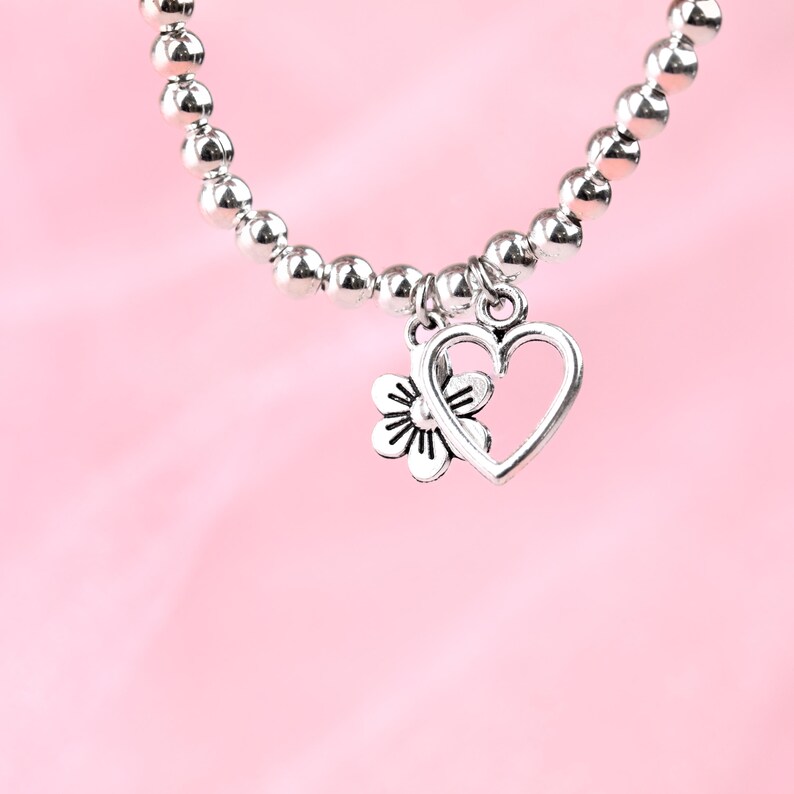 May include: A silver bracelet with a heart and flower charm. The bracelet is made of small silver beads and the charm is attached to the bracelet with a silver loop.