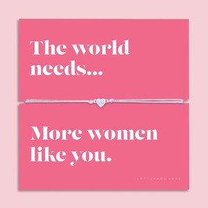 Op de afbeelding: Een roze kaart met de tekst "De wereld heeft... Meer vrouwen zoals jij nodig." Een zilveren armband met een hartvormige bedel is gecentreerd op de kaart. De woorden "LETTERBOX LOVE" staan onderaan.